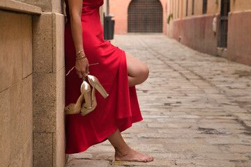 Woman in an elegant red party dress with golden high heels in her hand, barefoot, leaning against...