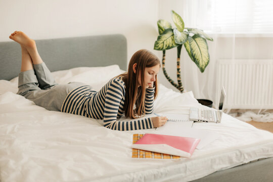 A charming teenage girl is lying on the bed, doing homework with a laptop, making notes in a notebook. The concept of training and education, modern technologies