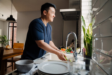 Adult brunette asian man wearing t-shirt washing dishes at kitchen