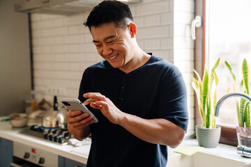 Adult brunette asian man smiling and using cellphone at home