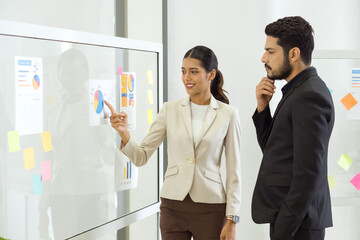 Young asian businesswoman in brown suit pointing finger at pie chart for business target, presented to investor. There was a note paper and document on the glass panel.