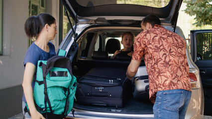 Family getting ready for summer field trip while loading vehicle with voyage trolleys and citybreak. Parents putting luggage in car trunk while unpatient daughter hurrying them.