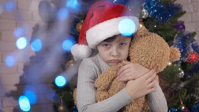 Upset Santa child with teddy bear. An upset Santa little girl stay with a teddy bear by the Christmas tree in the empty room.