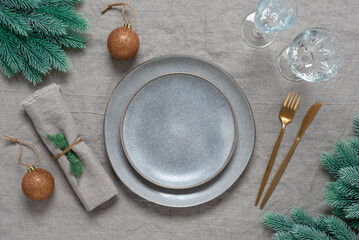 Christmas or New Year table setting. Empty mockup plates, golden cutlery, glasses, spruce branches and xmas decorations on a gray linen tablecloth. View from above, selective focus.