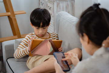 Fototapeta premium Asian boy sits on the sofa playing games with his tablet, with his single mother sitting next to him chatting with a friend on a mobile phone. A middle-aged man and woman sit in the living room