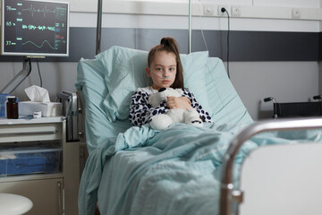 Ill kid resting in children healthcare facility patient bed alone while having teddy bear. Sick...