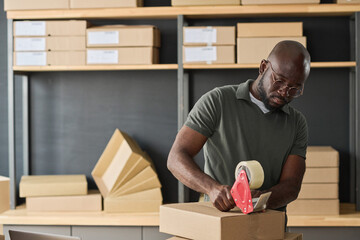 African warehouse worker in eyeglasses packing cardboard boxes with adhesive tape before shipping