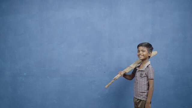 Smiling young boy kid holding cricket bat by looking at camera on blue background with copy space - concept of future goal, inspiration, childhood dream and poverty