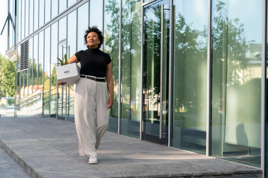 Smiling young businesswoman with box walking on footpath
