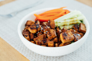 Bowl with roasted tempeh made of fermented soy beans, fresh vegetable sticks with sauce and sesame seeds on the table. Plant based protein. Healthy cooking and eating. Go vegan. selective focus