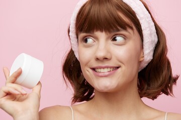 a happy woman stands on a pink background smiling happily and holding a white jar of cream in her hand looking away