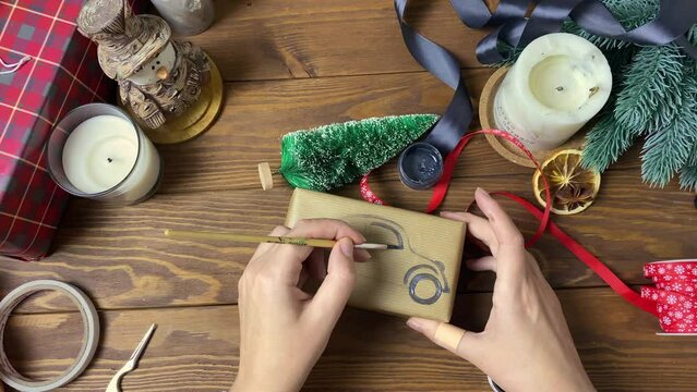 Women's hands draw car with brush and paint on gift box. Surprise in craft wrapper, decorative Christmas tree, candles, ribbons and snowman figure on wooden table. New Year mood.