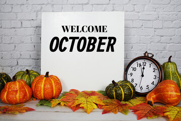 Welcome October text message with alarm clock, pumpkin and maple leaf decoration on white brick...