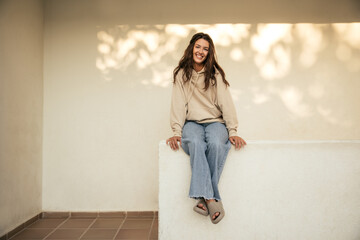 Pretty young caucasian brunette woman is resting sitting against white wall at sunset. Girl smiles, wears hoodie, jeans and slippers. Leisure lifestyle and beauty concept.