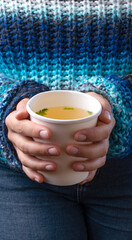 Close-up of Famele hands in blue sweater holding a Chicken broth soup in takeaway cup.