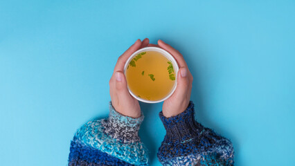 Famele hands holding a Chicken broth soup in takeaway cup. Blue background with copy space, Top...