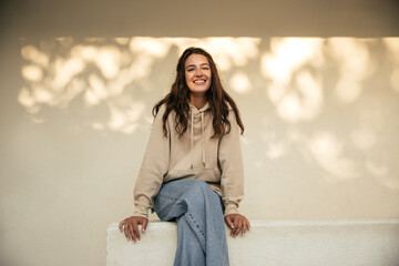 Happy young caucasian woman with snow-white smile sits on white wall background. Brunette wears...