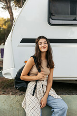 Calm young caucasian woman tourist looking at camera spends weekend outside city. Brunette wears tank top, jeans and backpack. Leisure lifestyle and beauty concept.