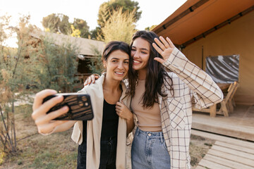 Cute young caucasian girls have photo session on smartphone outdoors. Brunettes smile, wear T-shirt, shirt, sweatshirt and pants. Concept of use