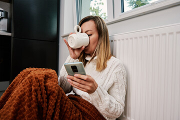 Worried sad woman sits under blanket near heating radiator and use smartphone, Rising costs in...