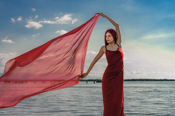 A beautiful graceful slender model girl in a silk or satin red dress with red hair poses and waves a red pareo handkerchief in the wind, against the background of the river and the sunset sky clouds - obrazy, fototapety, plakaty