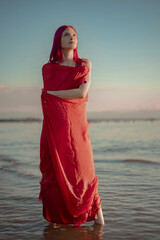 A beautiful slender girl with red hair stands in the river, posing for a photographer, wrapped in a red transparent cloth during sunset against the background of water and sky. Looks like monument