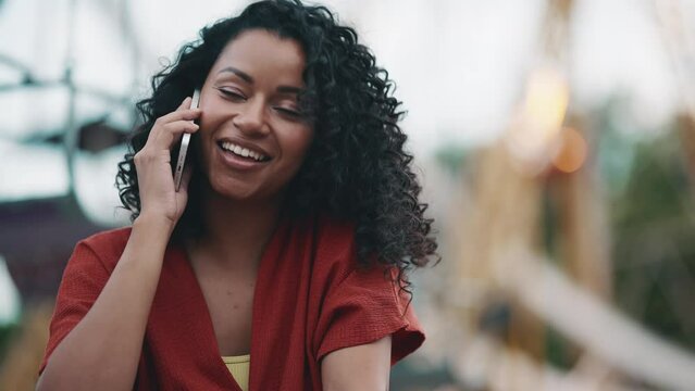 Smiling hispanic curly haired woman talking by phone while sitting in adventure park