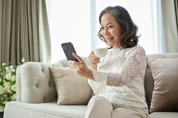 Gorgeous Asian-aged woman relaxes in the living room, sipping coffee, using smartphone