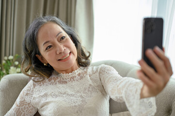 Happy Asian middle-aged woman uses her smartphone to take a selfie in her living room.