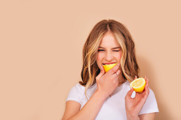 A teenage girl with wavy hair in a white t-shirt bites the lemon and grimaces in displeasure from...