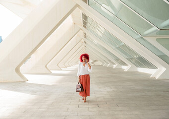 Fashionable and smiling Latin businesswoman with red afro hair talking on her smartphone while walking under a modern triangle-shaped architecture - Powered by Adobe
