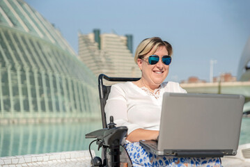 front view of a happy woman with disability with with sunglasses in wheelchair using laptop in...