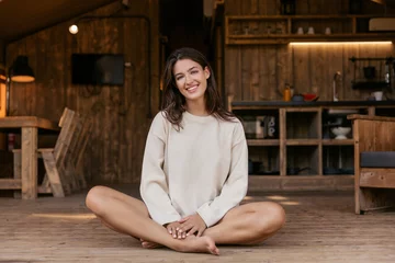 Tuinposter Attractive young caucasian woman smiling looking at camera sitting in lotus position indoors. Brunette wears casual spring clothes. Lifestyle concept © Look!