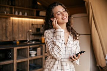 Fototapeta na wymiar Tender young caucasian woman listens to music using headphones and phone standing indoors. Brunette, closing her eyes, wears shirt. Online entertainment concept