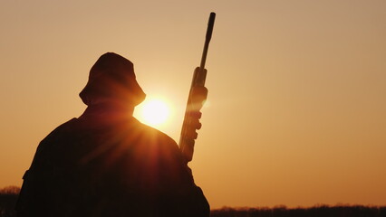A hunter with a gun goes forward across the field towards the setting sun. Hobby for men