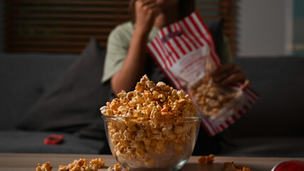 Bowl of popcorn and on wooden table with young woman watching television on couch in background. Relaxation and hobby concept