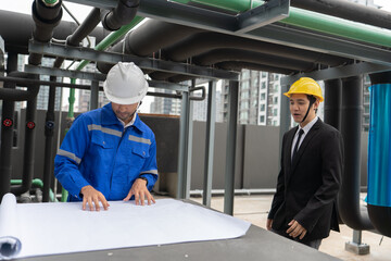 At construction site manager visit engineer working on the rooftop near pipe rack to inspection the progress by looking from the blue print drawing and look around and pointing and check hand
