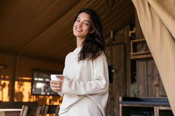Positive young caucasian brunette woman smiles teeth looking at camera relaxing in camping. Girl with cup of tea wears white casual clothes. Lifestyle, different emotions, leisure concept