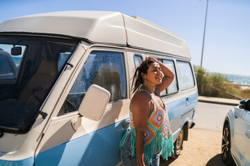 Chica joven guapa posando y sonriendo delante de furgoneta vintage en aparcamiento de la playa