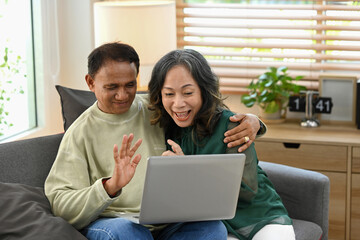 Happy retired couple having fun web surfing internet or making video call with family or friends on laptop together at home