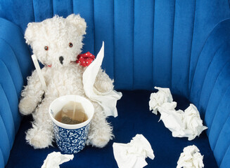 sick teddy bear with fever thermometer, hot tea and a lot of handkerchiefs on blue velvet armchair...