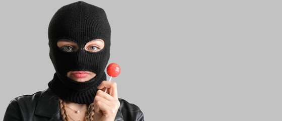 Young woman in balaclava and with lollipop on grey background with space for text