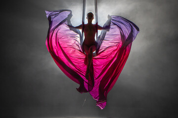 Silhouette of a young woman balancing on an airy silk ribbon and waving a cloth like butterfly wings. A female athlete performing in a circus at a height in the dark with backlight. - obrazy, fototapety, plakaty