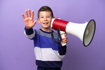 Little boy isolated on purple background holding a megaphone and saluting with hand with happy...