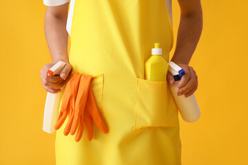 Young man in apron with cleaning supplies on yellow background, closeup