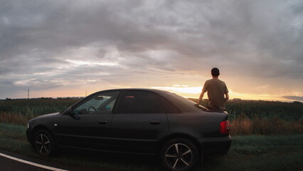 A young man is resting from the road, sitting on the trunk of his car and enjoying the colors of the sunset in cloudy rainy weather. Traveling by car concept