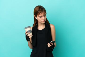 Redhead sport girl isolated on blue background holding coffee to take away and a mobile