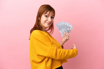 Redhead girl taking a lot of money isolated on pink background pointing back