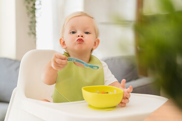 Adorable blond baby boy sitting at table in high chair with spoon in hand and yellow plate with...
