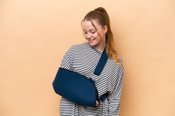 Young caucasian girl with broken arm and wearing a sling isolated on beige background with happy...